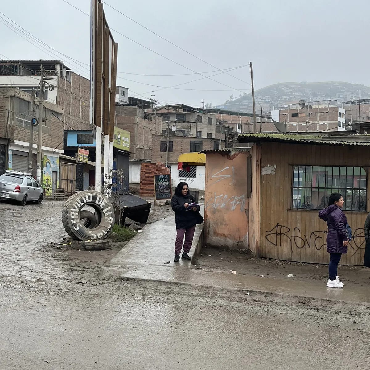 Calle sin asfaltar de un barrio en ladera: la socia camina con sus papeles en la mano entre viviendas y bodegas